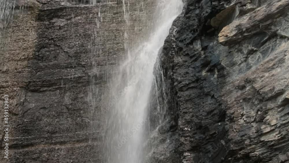 The camera tilts up as water cascades down a steep rocky cliff with green bushes growing from a ledge near the top of the waterfall. The camera tilts up as water cascades down a steep rocky cliff with green bushes growing from a ledge near the top of the waterfall.