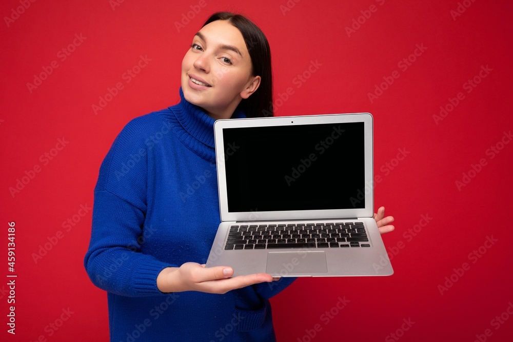 Charming pretty happy beautiful young brunet female student lady holding computer laptop looking at camera in blue sweater isolated over red wall background Charming pretty happy beautiful young brunet female student lady holding computer laptop looking at camera in blue sweater isolated over red wall background