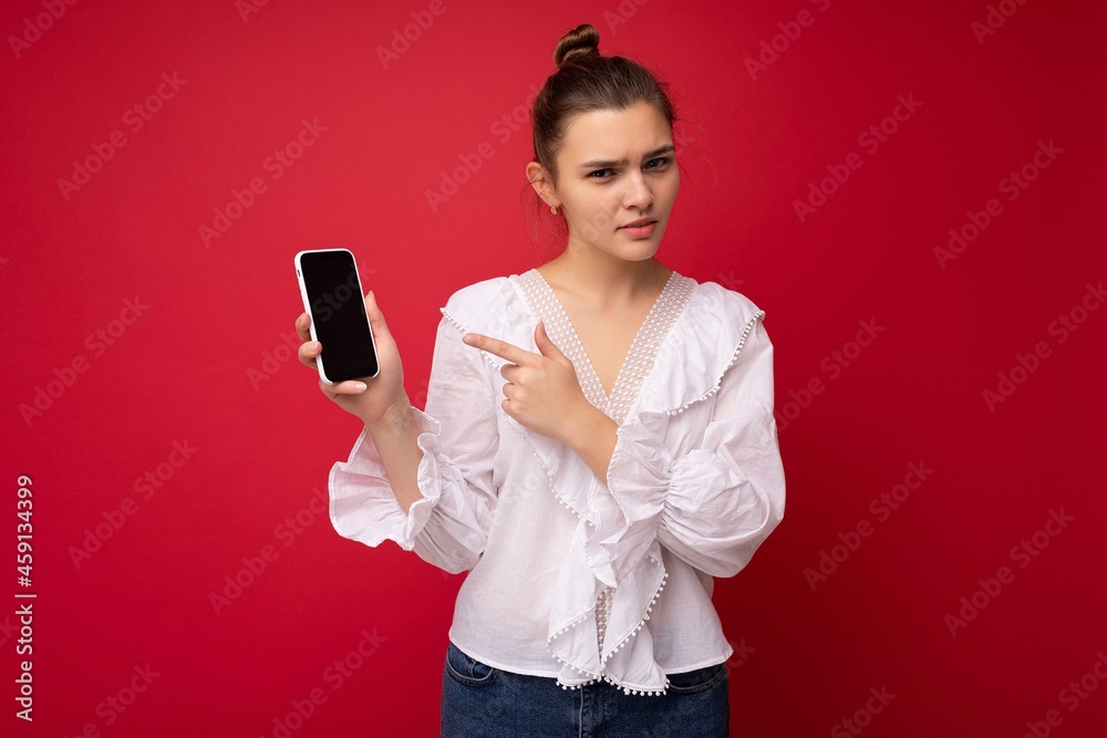 Attractive young dissatisfied brunet woman wearing white blouse standing isolated over red background showing mobile phone with empty screen for cutout looking at camera and having troubles Attractive young dissatisfied brunet woman wearing white blouse standing isolated over red background showing mobile phone with empty screen for cutout looking at camera and having troubles