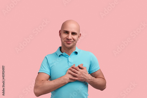 Believe me. Sincere bald homosexual man with bristle falls in love, presses hands to heart, keeps palms on chest, smiles, tilts head, gay friendly, wears blue polo shirt, stands over pink background.