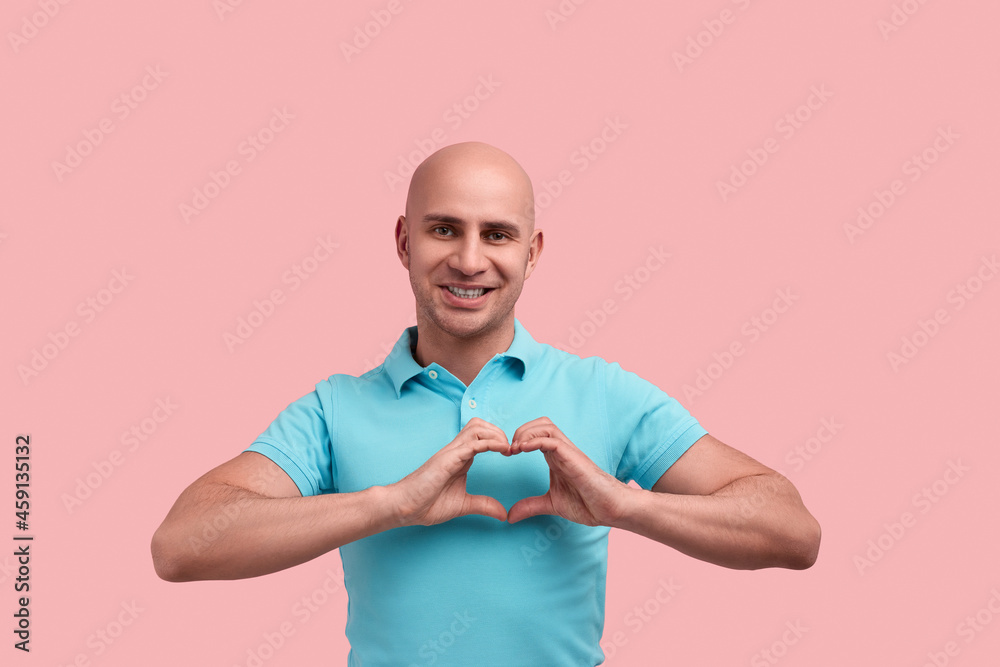 Be my valentine. Smiling bald homosexual man with bristle makes heart gesture with hands against chest, falls in love, looks straight, gay friendly, wears blue polo shirt, stands over pink background.