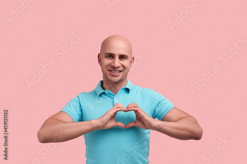 Be my valentine. Smiling bald homosexual man with bristle makes heart gesture with hands against chest, falls in love, looks straight, gay friendly, wears blue polo shirt, stands over pink background.