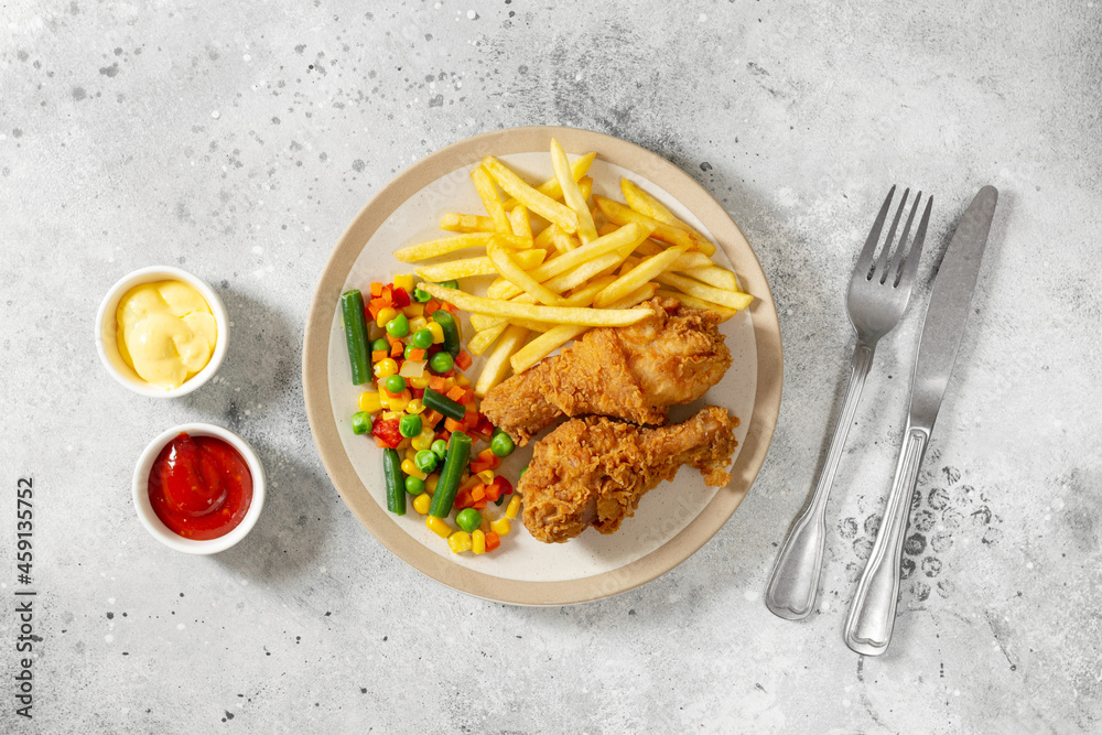Fried crispy chicken legs in breadcrumbs with french fries and vegetables in a plate on a culinary background. Fast food from the drumstick top view