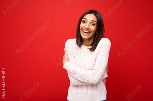 Photo of young beautiful happy smiling brunette woman wearing trendy light jersey . Sexy carefree female person posing isolated near red wall in studio with free space. Positive model with natural
