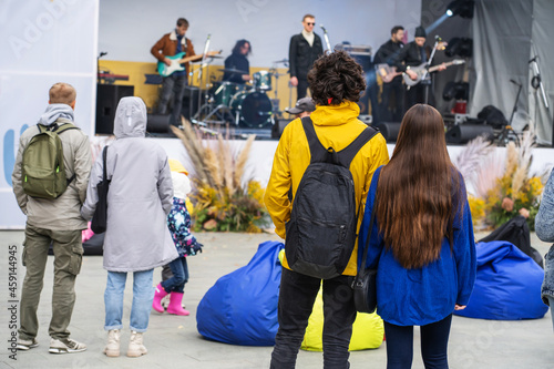 Young couple in warm jackets at a music concert in a city park. People watch the performance of a rock band on the street stage. Unrecognizable people. Autumn season
