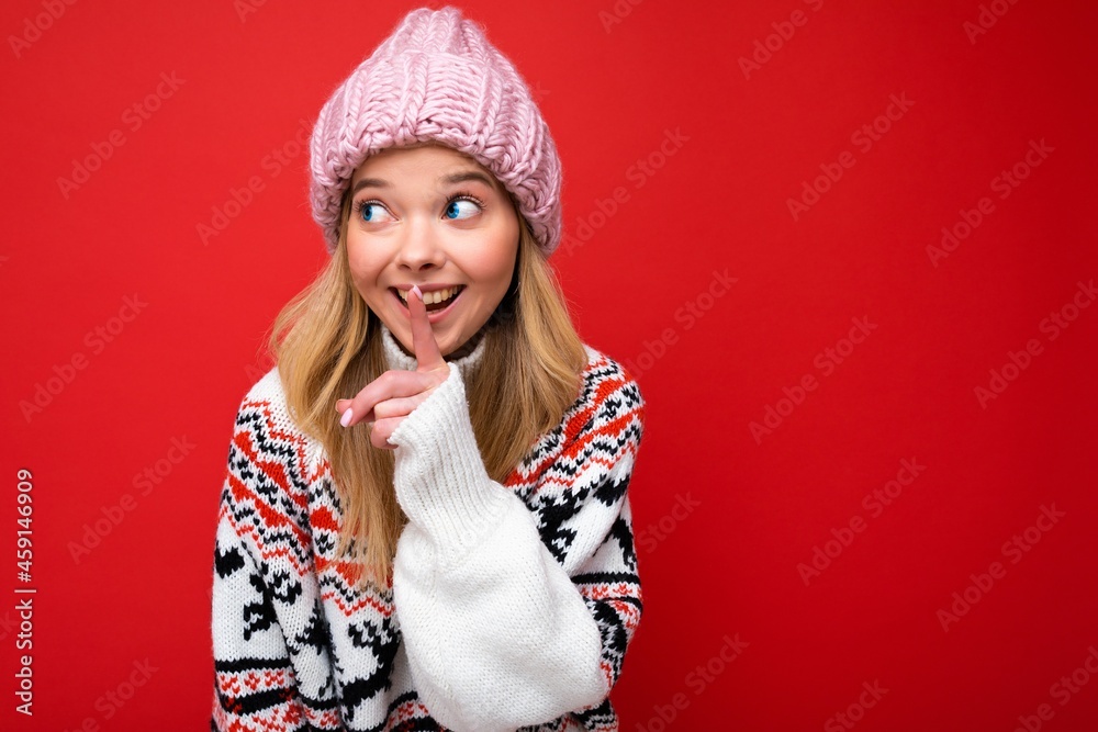 Portrait of young positive happy attractive blonde woman with sincere emotions wearing pink hat and winter sweater isolated over red background with copy space and asking remain silent. Be quiet Portrait of young positive happy attractive blonde woman with sincere emotions wearing pink hat and winter sweater isolated over red background with copy space and asking remain silent. Be quiet