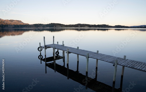 Magical view of the wooden dock in the placid lake at sunrise. The mountains, forest and blue sky reflection in the water.