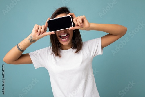 Photo of pretty positive young brunette female peson good looking wearing white t-shirt standing isolated on blue background with copy space holding smartphone showing phone in hand with empty screen photo