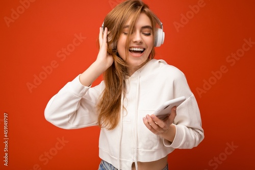 Photo of beautiful joyful smiling young woman wearing stylish casual clothes isolated over background wall holding and using mobile phone wearing white bluetooth headphones listening to music and photo