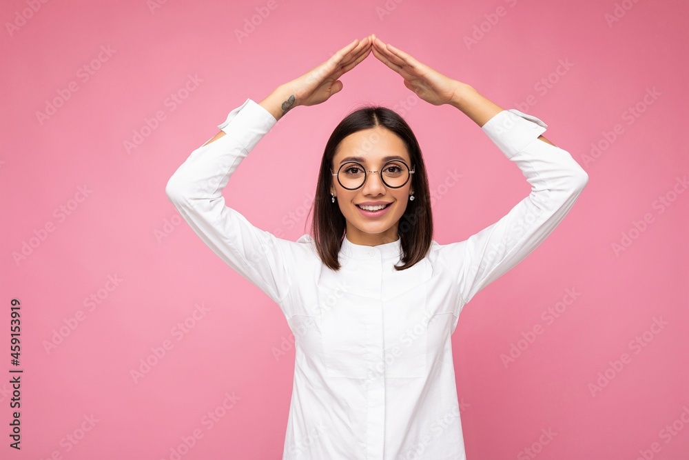 Shot of pretty positive smiling young brunette woman wearing white shirt and stylish optical glasses isolated over pink background looking at camera and showing home gesture sign Shot of pretty positive smiling young brunette woman wearing white shirt and stylish optical glasses isolated over pink background looking at camera and showing home gesture sign