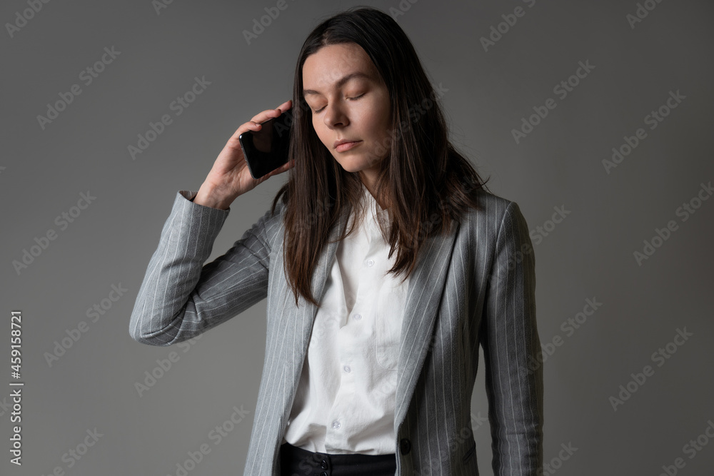 businesswoman dressed in a jacket and trousers in a casual style uses a smartphone for work and contact with partners, studio portrait on a gray, phone conversation, calmly listen to the interlocutor businesswoman dressed in a jacket and trousers in a casual style uses a smartphone for work and contact with partners, studio portrait on a gray, phone conversation, calmly listen to the interlocutor