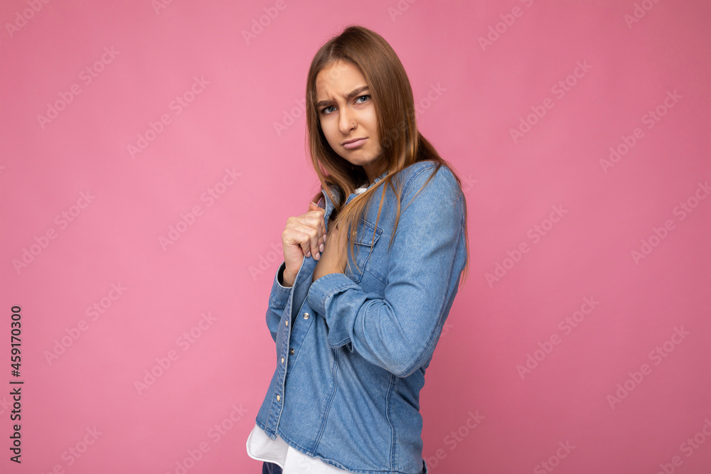 Portrait of sad dissatisfied angry young beautiful dark blond woman with sincere emotions wearing blue denim shirt isolated over pink background with copy space and hiding. Negative concept Portrait of sad dissatisfied angry young beautiful dark blond woman with sincere emotions wearing blue denim shirt isolated over pink background with copy space and hiding. Negative concept