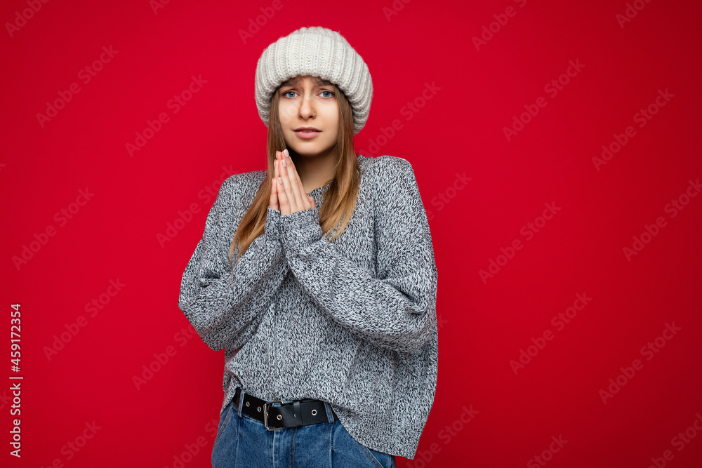 Portrait of sad asking young beautiful dark blonde woman with sincere emotions wearing grey pullover and beige knitted hat isolated over red background with empty space and holding hands in pray Portrait of sad asking young beautiful dark blonde woman with sincere emotions wearing grey pullover and beige knitted hat isolated over red background with empty space and holding hands in pray