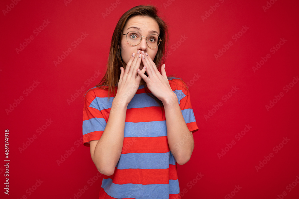 Photo of young cool attractive brunette woman with sincere emotions wearing trendy striped t-shirt and optical glasses isolated over red background with free space and covering mouth Photo of young cool attractive brunette woman with sincere emotions wearing trendy striped t-shirt and optical glasses isolated over red background with free space and covering mouth