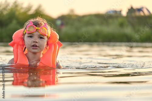 A little girl bathes in the water in the sunset light. Child playing on the beach during summer vacation. The child swims in a life jacket and goggles. Safety on the water.