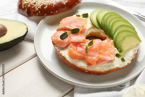 Delicious bagel with cream cheese, salmon and avocado on white wooden table, closeup