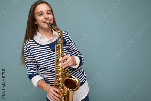 Photo shot of beautiful happy smiling brunette little girl wearing striped longsleeve standing isolated over blue background wall playing saxophone looking at camera photo