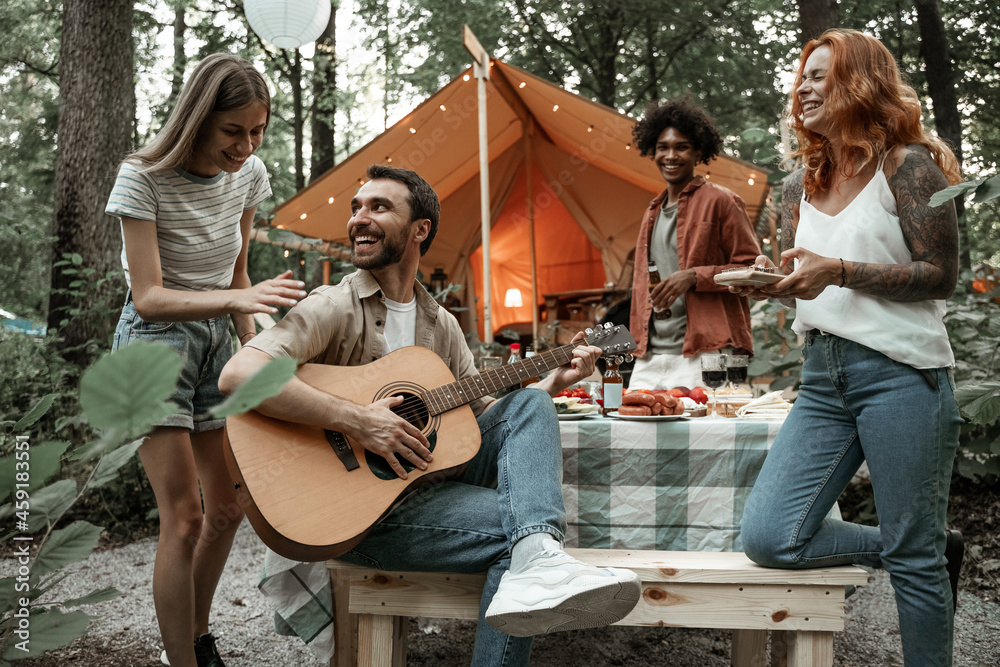 Group of young friends traveling in glamping in the forest having fun playing guitar and mbira kalimba roasting sausages sitting at dinner table near tent during summer vacation laughing hanging out Group of young friends traveling in glamping in the forest having fun playing guitar and mbira kalimba roasting sausages sitting at dinner table near tent during summer vacation laughing hanging out
