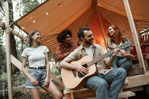 Group of young friends traveling in glamping in the forest having fun playing guitar  mbira or kalimba and singing songs roasting sausages sitting at tent during summer vacation laughing hanging out