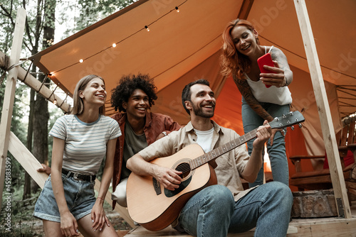 Close up portrait of group of young friends hanging out traveling together in glamping in forest having fun playing guitar, making selfie, streaming to social media sitting in tent laughing