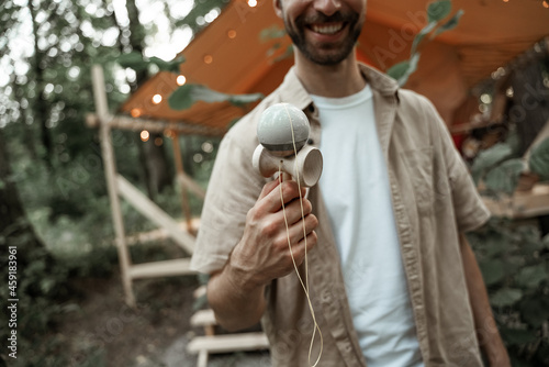 Young smiling man with stubble enjoy playing kendama at camping site, traditional skill toy. Cup-and-ball game. Wooden toy. Millennial with japanees toy entertaining friends in summer glamping