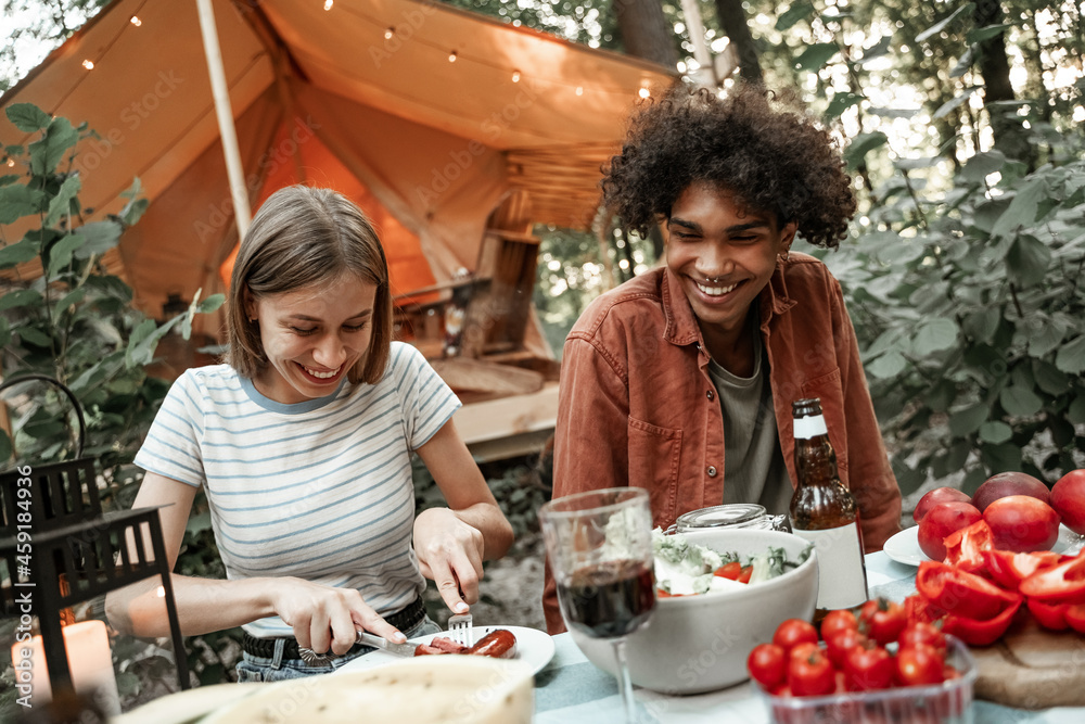 Young multiracial couple having dinner at glamping, laughing after sunset. Happy millennials camping at open air picnic under bulb lights. Spending time with friends outdoors, barbeque party