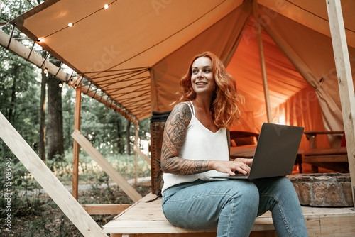 Red-haired young woman with tattoo working remotely on laptop at glamping. Camping lifestyle vacation. Low budget travel, holiday in forest. Wi-fi connection information communication technology.
