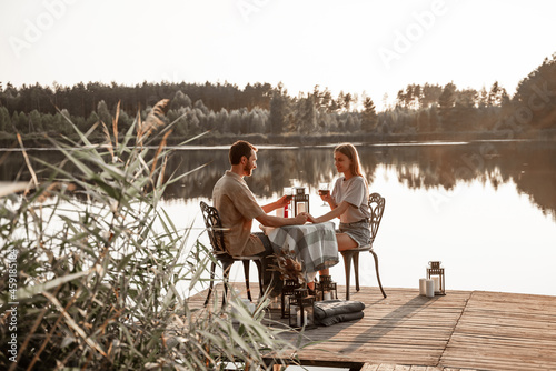 Young couple sitting at the table spend time together on wooden pier on forest lake celebrating anniversary, holding hands. Love is in the air, love story concept. Romantic date on lake with candles.
