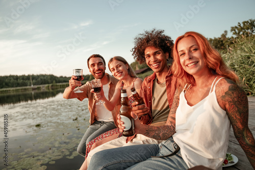 Group of friends having fun on picnic near a lake, sitting on wooden pier eating and drinking wine, beer, cider. Smiling young people having party celebration outdoors during sunset in countryside