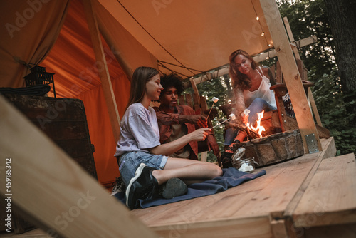 Group of young smiling people roasting marshmallows on skewers over fire pit at campsite, enjoying outdoor glamping holiday with friends togetherness reopen after pandemic lockdown.