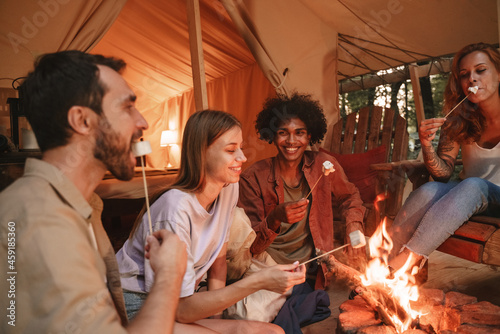 Group of young smiling people roasting marshmallows on skewers over fire pit at campsite, enjoying outdoor glamping holiday with friends togetherness reopen after pandemic lockdown.