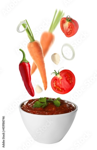 Different ingredients falling into ceramic bowl with delicious adjika sauce on white background