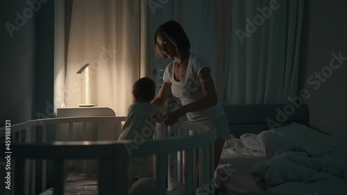 Night shot of a Mother waking up at night to a crying Baby in Child Crib. Caucasian Neonate Toddler at Home in Kids Bedroom. Concept of Childhood, New Llife and Parenthood.