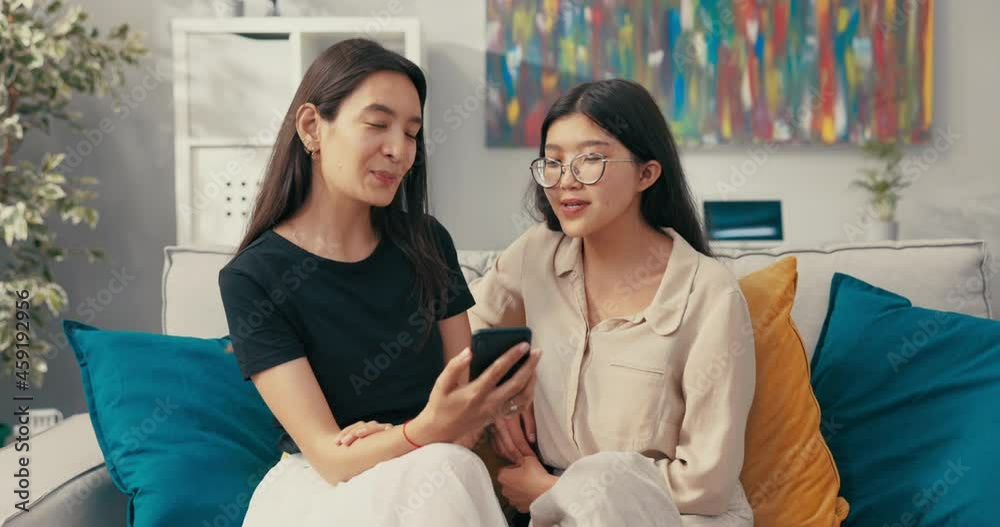 Two pretty best friends of Asian Korean beauty spend an afternoon on the living room couch, talking, the woman shows vacation photos on her phone, they look together, smile, comment Two pretty best friends of Asian Korean beauty spend an afternoon on the living room couch, talking, the woman shows vacation photos on her phone, they look together, smile, comment