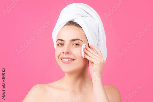 Beautiful girl with a towel on head clean remove cosmetics from face use cotton patch isolated over pink background. Facial treatment, cosmetology, beauty and spa.