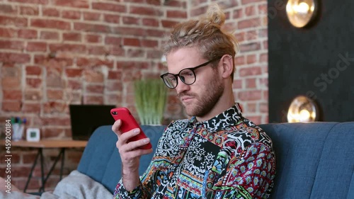 Young focused male in glasses sitting on couch tapping on mobile phone. Gorgeous man texting on smartphone, view photos on the phone. Serious guy using smartphone at home. Technology concept.