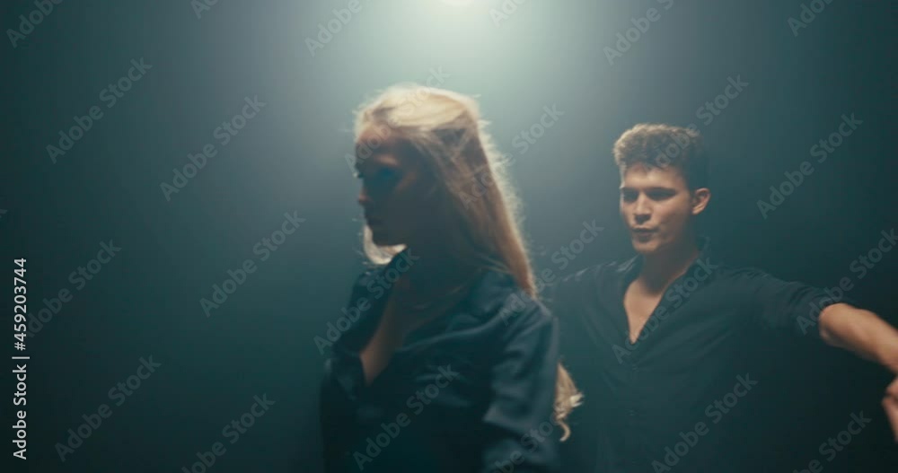 Prosfessional pair of dancers hang out in dark studio for dance practice, man in black shirt holds back partner who turns around approaches guy touching shirt, close zone between couple Prosfessional pair of dancers hang out in dark studio for dance practice, man in black shirt holds back partner who turns around approaches guy touching shirt, close zone between couple