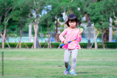 Cute girl is walking, in her hands she holds a badminton racket ball. Child is feeling embarrassed. Natural background and green lawn. Children aged 4-5 years old.