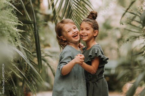 Obraz na plátně Two cute smiling little girls belonging to different races, in linen clothes, holding hands and walking in the botanical garden