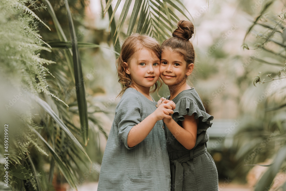 Naklejka premium Two cute smiling little girls belonging to different races, in linen clothes, holding hands and walking in the botanical garden. Children explore tropical plants and flowers in the greenhouse.