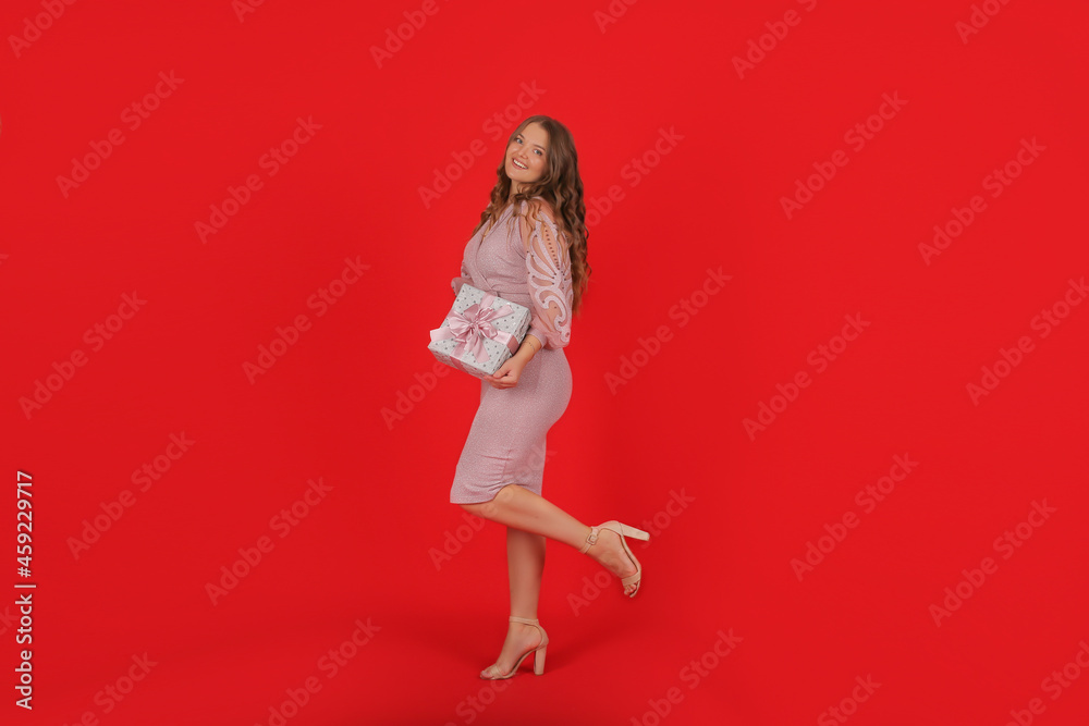 a young happy beautiful elegant girl with long hair in a pink dress on a red background holds a holiday gift box in her hands a young happy beautiful elegant girl with long hair in a pink dress on a red background holds a holiday gift box in her hands