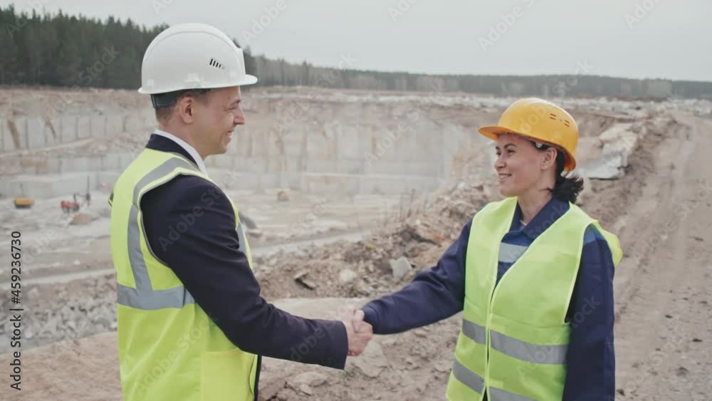 Slowmo tracking shot of happy male manager and female supervisor in safety gear standing on top of granite quarry and shaking hands while chatting and laughing