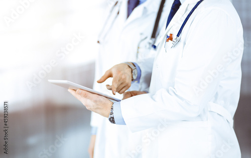 Two unknown doctors with stethoscopes discussing medical exam resoults, standing at sunny hospital office. Physicians use a computer tablet for filling up medication names records. Perfect medical