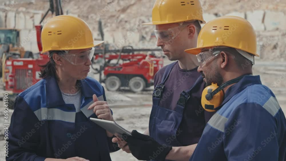 Handheld tracking of female supervisor in hard hat and safety goggles pointing at something on tablet and instructing male colleagues in granite quarry