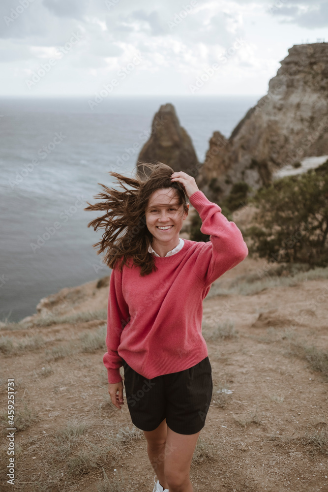 a young smiling brunette woman stands against the background of the sea and rocks in a red sweater. a traveler in a nature reserve