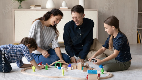 Happy young hispanic couple parents having fun with little adorable kids son daughter playing wooden toys railway road, sitting together on floor carpet, joyful family entertaining in living room.
