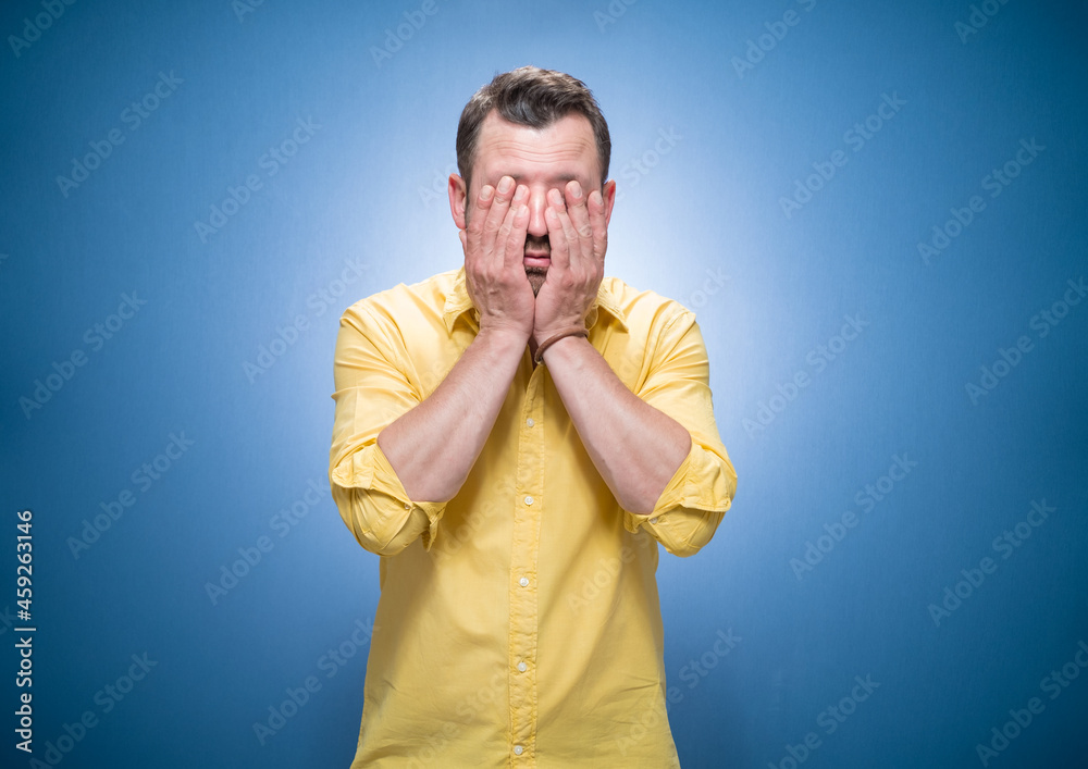 Tired guy over blue background, dresses in yellow shirt. Sleepy man rubbing her eyes. Young man covers face. Sleepy expression Tired guy over blue background, dresses in yellow shirt. Sleepy man rubbing her eyes. Young man covers face. Sleepy expression