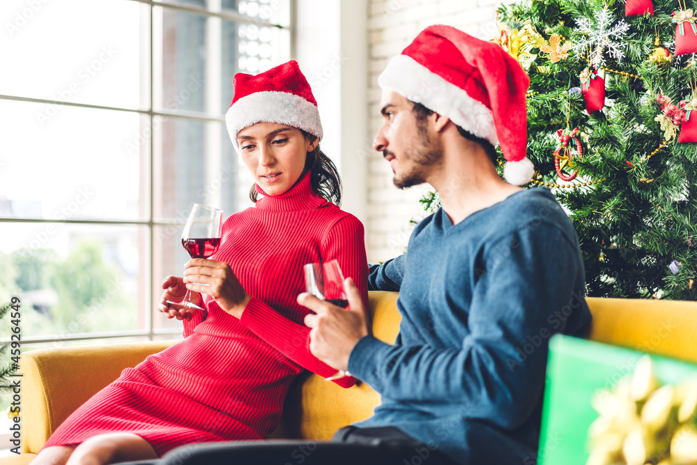 Fototapeta premium Romantic sweet couple in santa hats having fun and drinking wine glasses while celebrating new year eve and enjoying spending time together in christmas time at home