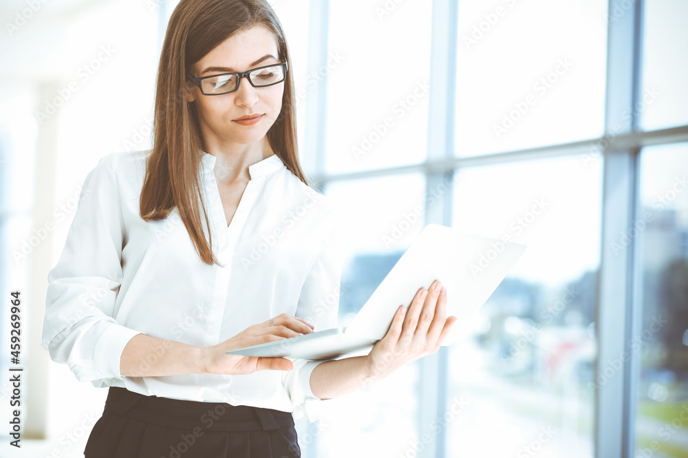 Beautiful female specialist with laptop computer standing in modern office and smiling charmingly. Working on design, data analysis, plan strategy. Business people concept Beautiful female specialist with laptop computer standing in modern office and smiling charmingly. Working on design, data analysis, plan strategy. Business people concept