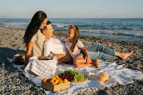 Vegan family spending time together outdoor. Picnic lunch with fruits by the seaside. Happy people eating natural healthy food, drinking juice and sitting on blanket on the beach.